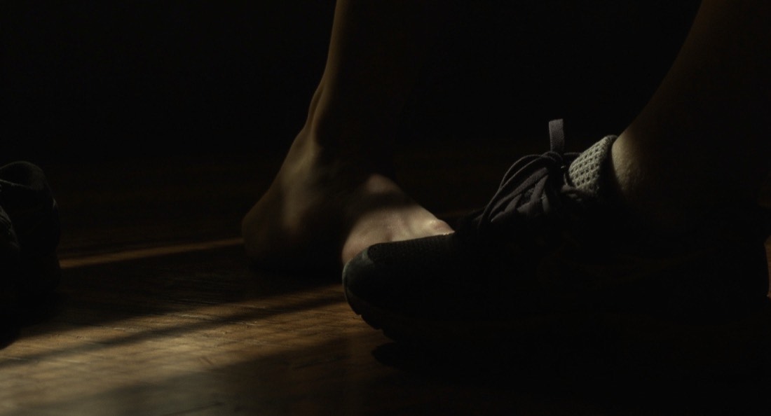 Bare feet on a dark wooden floor beside a pair of worn running shoes
