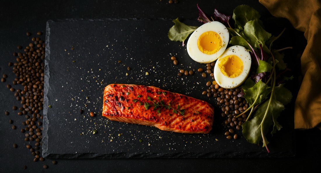 High-protein whole foods arranged on dark slate: salmon, eggs, yogurt, lentils and greens